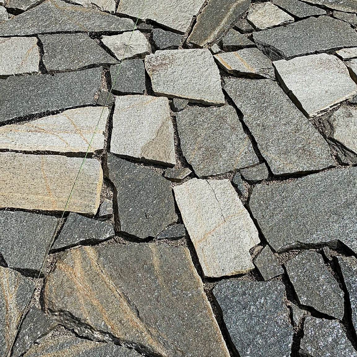 Bodenbeläge aus Naturstein mit unregelmäßig geformten Steinen in kühlen Grautönen. Die Steine variieren von hellem bis dunklem Grau, mit vereinzelten bläulichen und grünlichen Nuancen. Die raue und strukturierte Oberfläche der Steine, kombiniert mit den breiten und ungleichmäßigen Fugen, verleiht dem Belag eine rustikale und natürliche Optik.